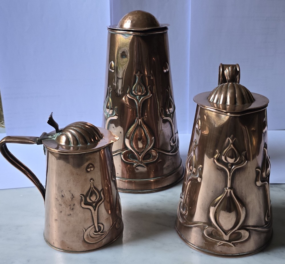 A set of three graduated copper jugs featuring embossed Art Nouveau floral motifs and shell-shaped hinged lids. View VII