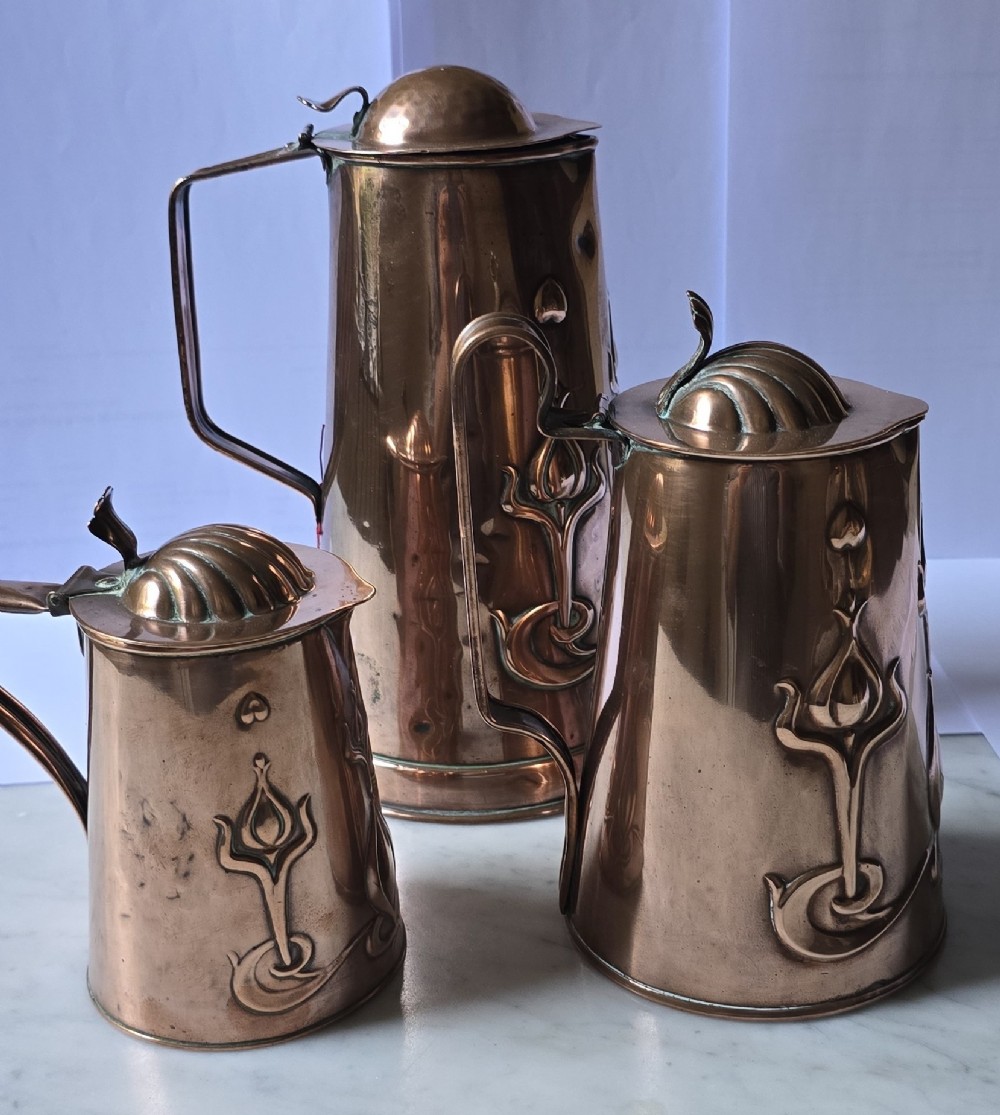 A set of three graduated copper jugs featuring embossed Art Nouveau floral motifs and shell-shaped hinged lids. View IX
