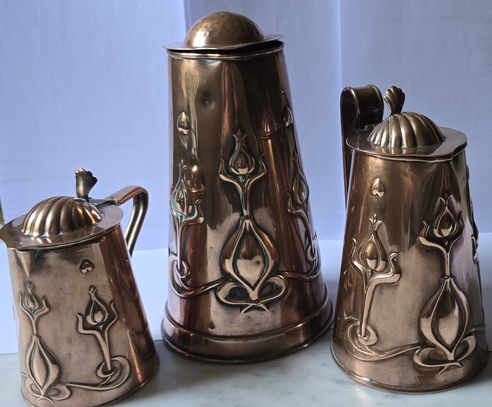 A set of three graduated copper jugs featuring embossed Art Nouveau floral motifs and shell-shaped hinged lids. View XIII