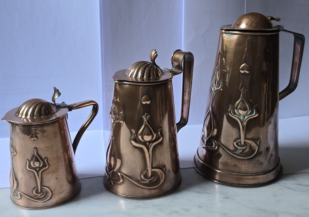 A set of three graduated copper jugs featuring embossed Art Nouveau floral motifs and shell-shaped hinged lids. View XIV