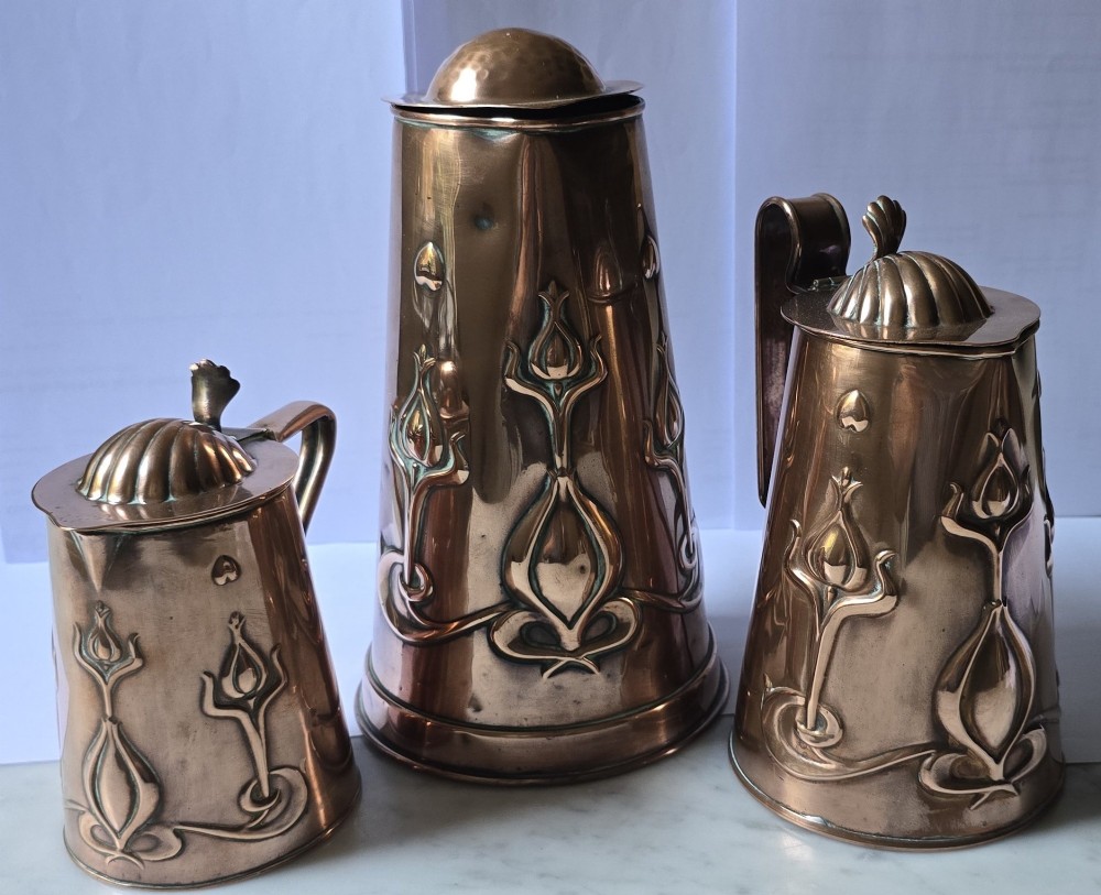 A set of three graduated copper jugs featuring embossed Art Nouveau floral motifs and shell-shaped hinged lids. View XV