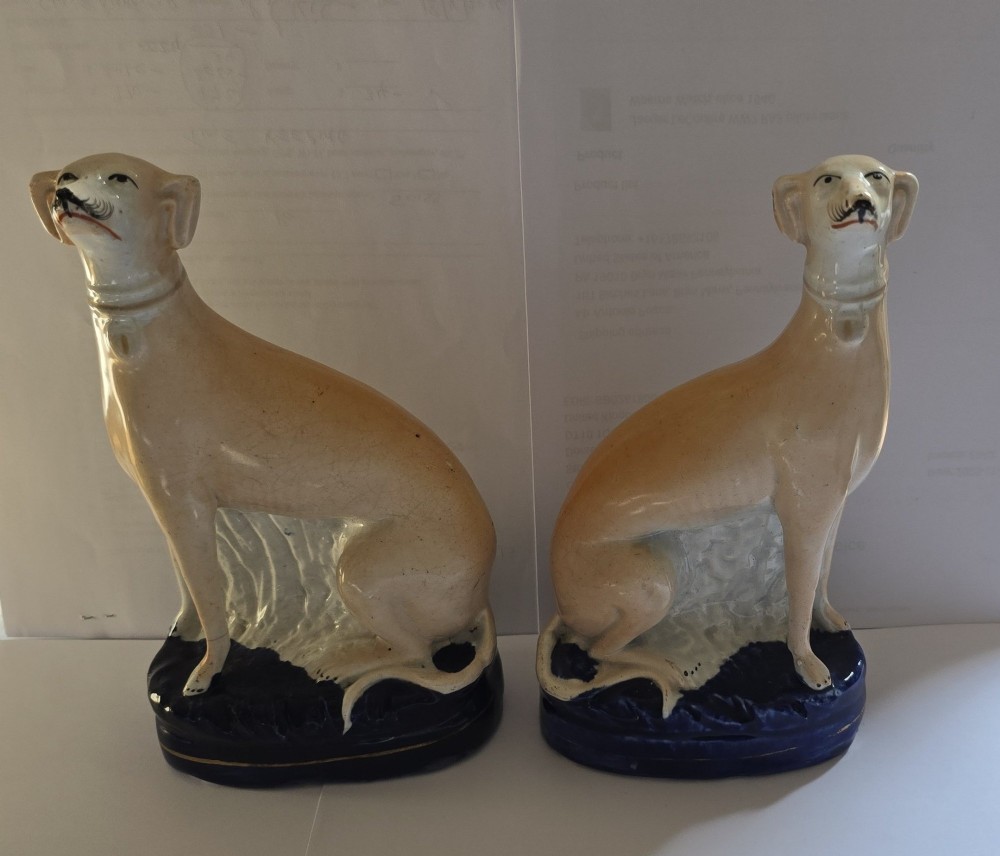 A pair of Victorian Staffordshire earthenware figurines depicting seated buff-coloured whippet dogs positioned on cobalt blue bases.