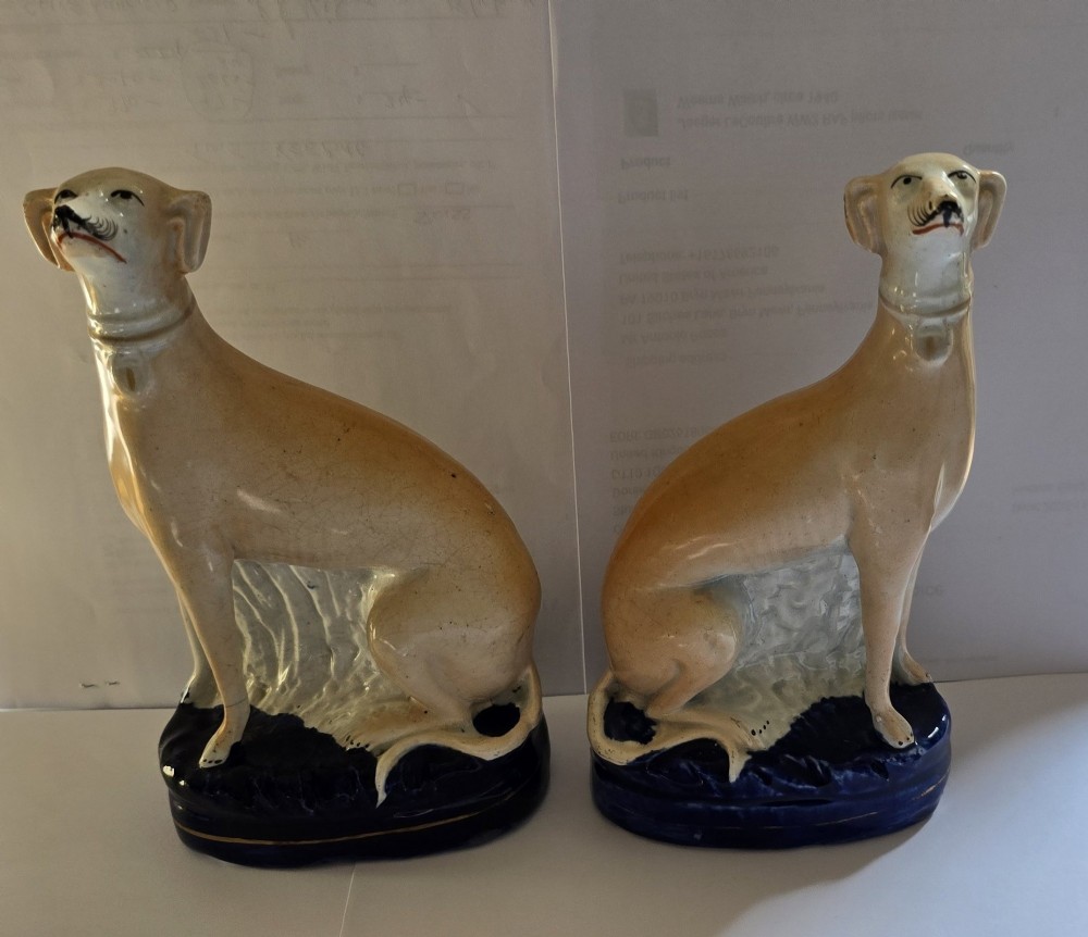 A pair of Victorian Staffordshire earthenware figurines depicting seated buff-coloured whippet dogs positioned on cobalt blue bases. View II