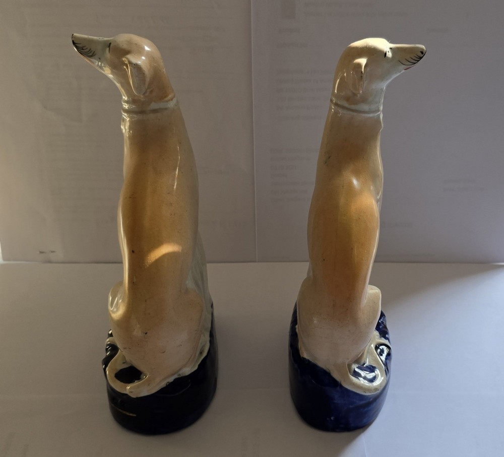 A pair of Victorian Staffordshire earthenware figurines depicting seated buff-coloured whippet dogs positioned on cobalt blue bases. View III