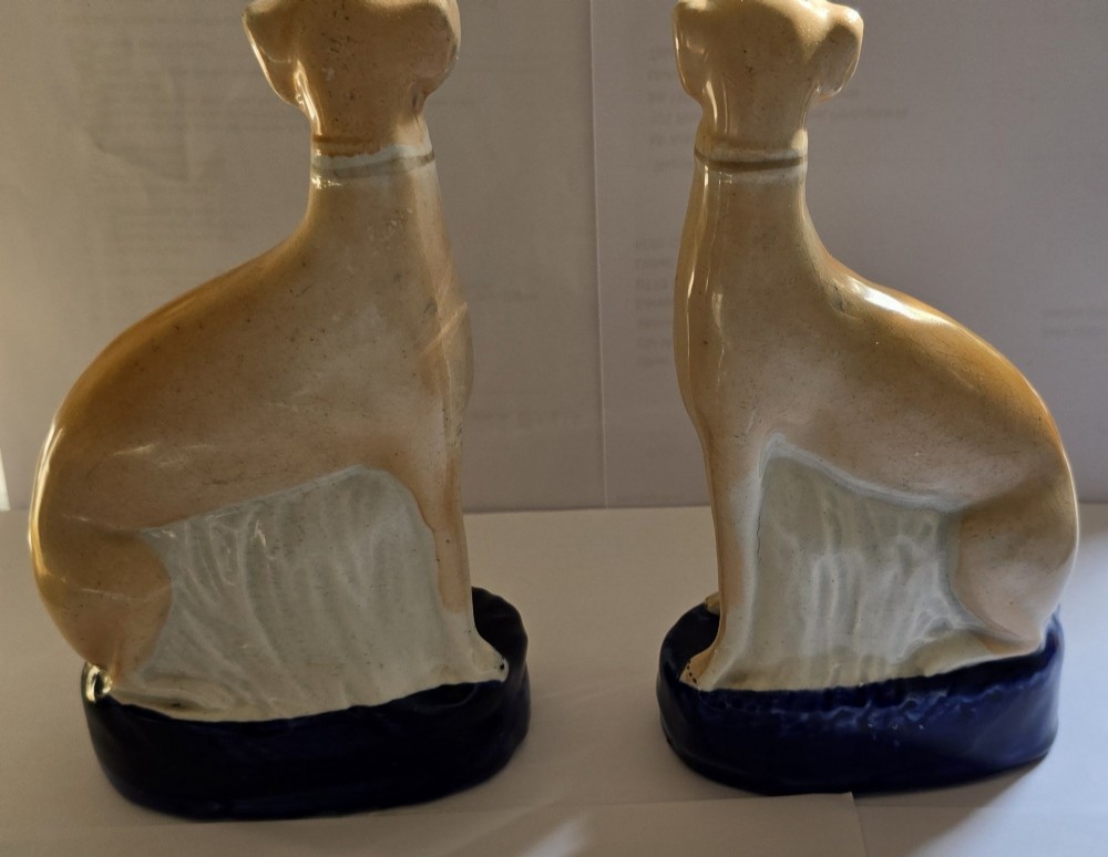A pair of Victorian Staffordshire earthenware figurines depicting seated buff-coloured whippet dogs positioned on cobalt blue bases. View VII