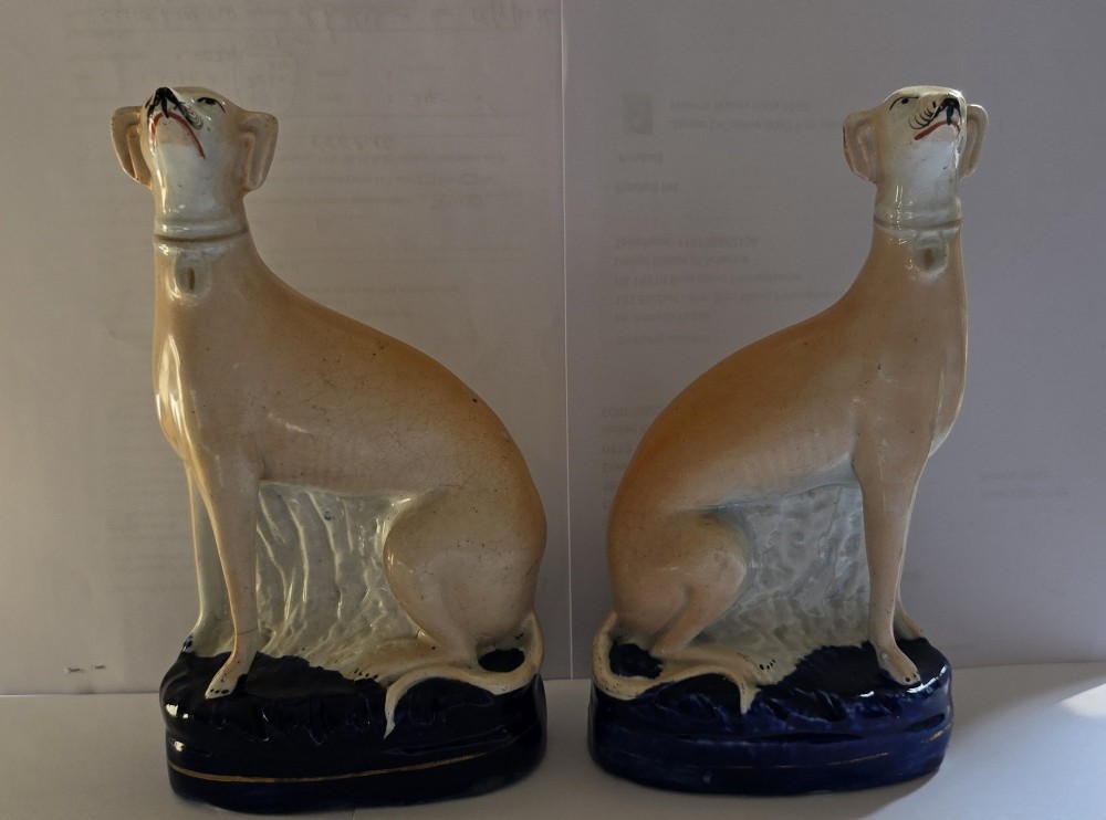 A pair of Victorian Staffordshire earthenware figurines depicting seated buff-coloured whippet dogs positioned on cobalt blue bases. View XII