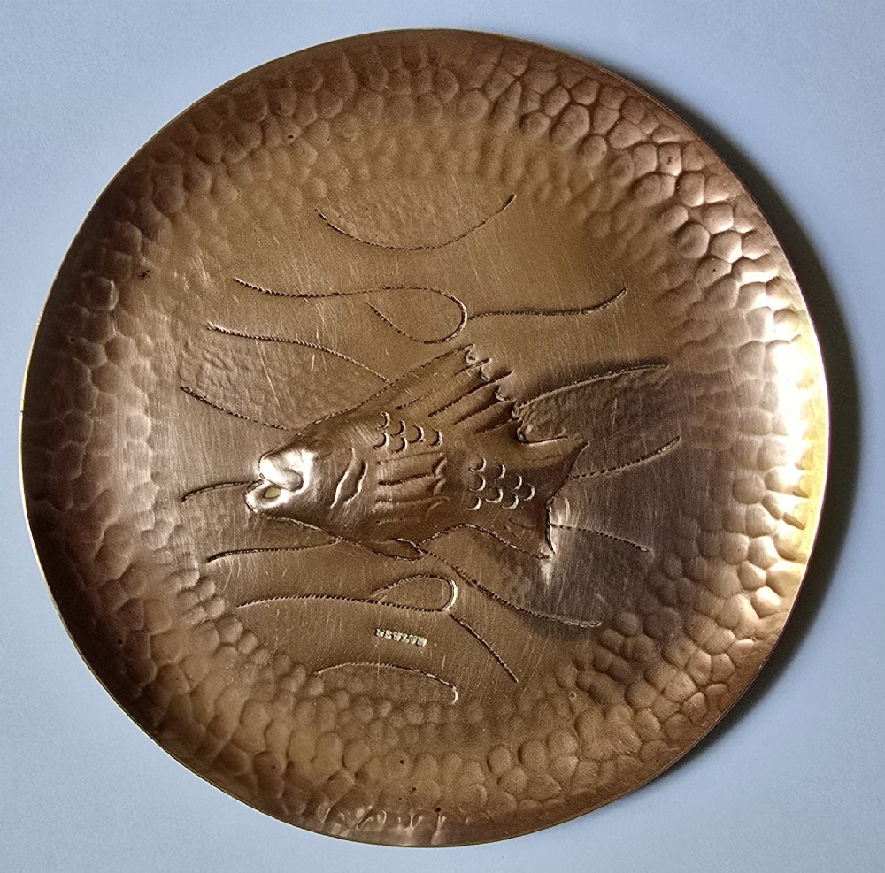 A circular hand-hammered copper visiting card tray featuring a central repousse fish motif surrounded by chased water lines and a spot-hammered rim.