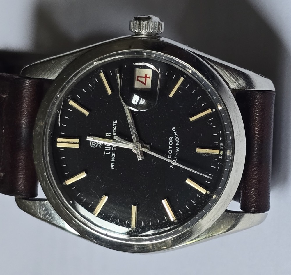 A vintage Tudor Prince Oysterdate wristwatch with a stainless steel case, a black dial featuring baton hour markers, a date aperture at three oclock, and a dark brown leather strap.