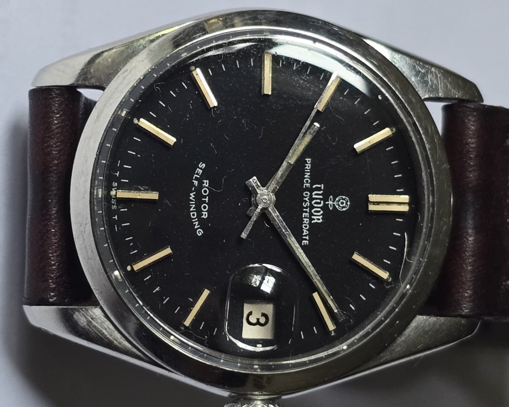 A vintage Tudor Prince Oysterdate wristwatch with a stainless steel case, a black dial featuring baton hour markers, a date aperture at three oclock, and a dark brown leather strap. View II
