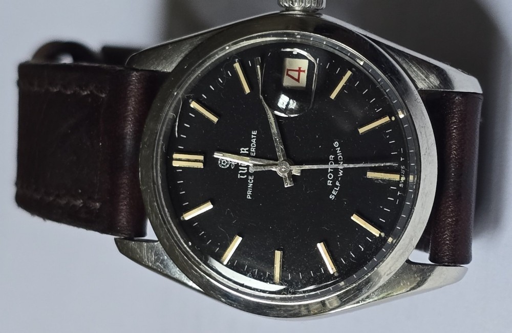 A vintage Tudor Prince Oysterdate wristwatch with a stainless steel case, a black dial featuring baton hour markers, a date aperture at three oclock, and a dark brown leather strap. View III