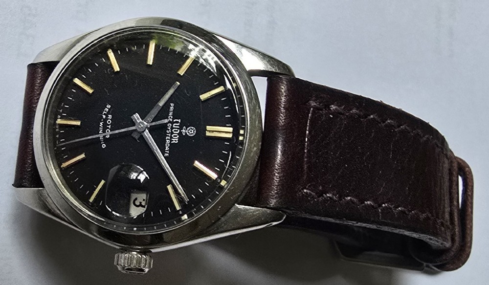 A vintage Tudor Prince Oysterdate wristwatch with a stainless steel case, a black dial featuring baton hour markers, a date aperture at three oclock, and a dark brown leather strap. View IV
