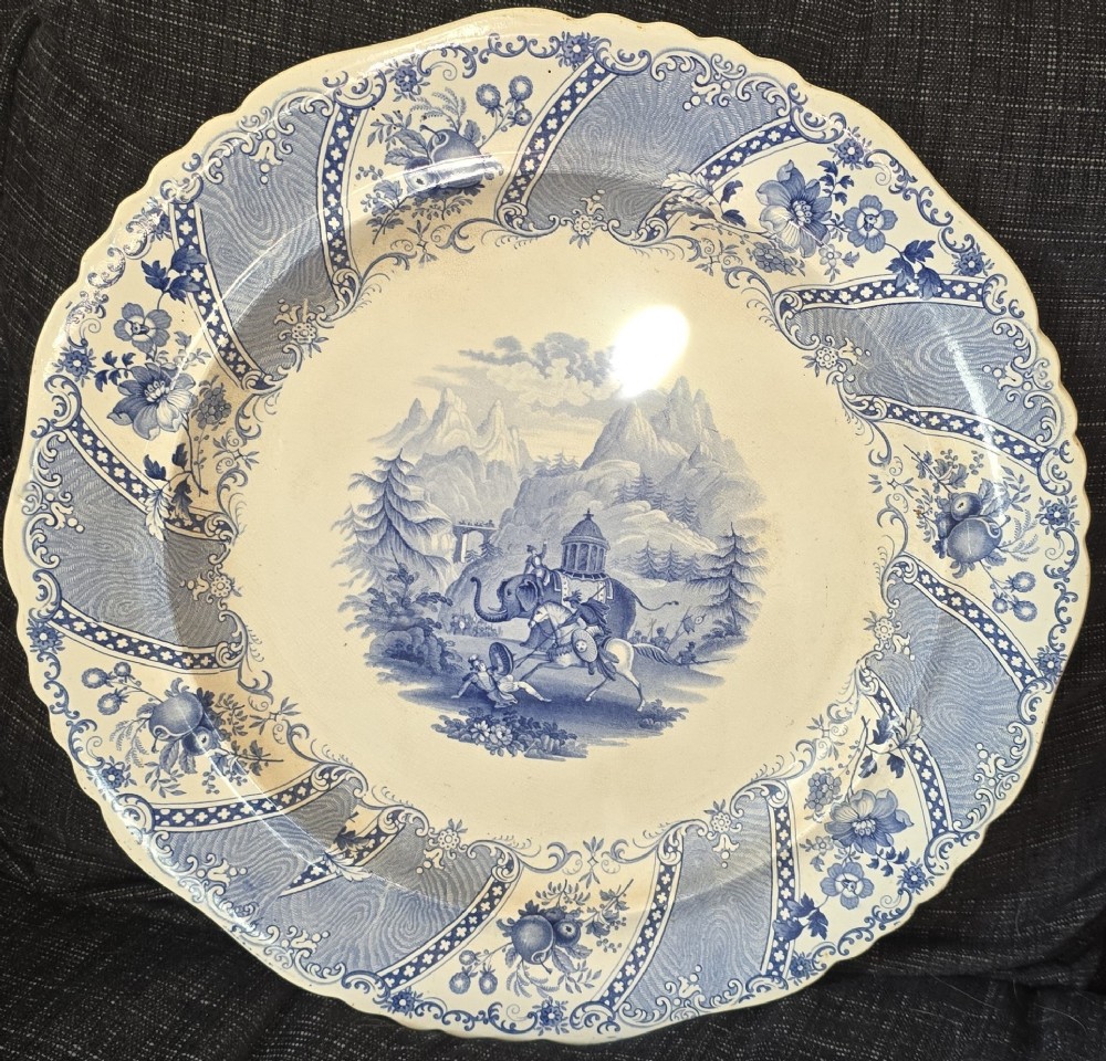 An antique circular earthenware plate featuring a blue transfer-printed central scene of Hannibal with an elephant and figures in a mountainous landscape, surrounded by an ornate scalloped floral border.