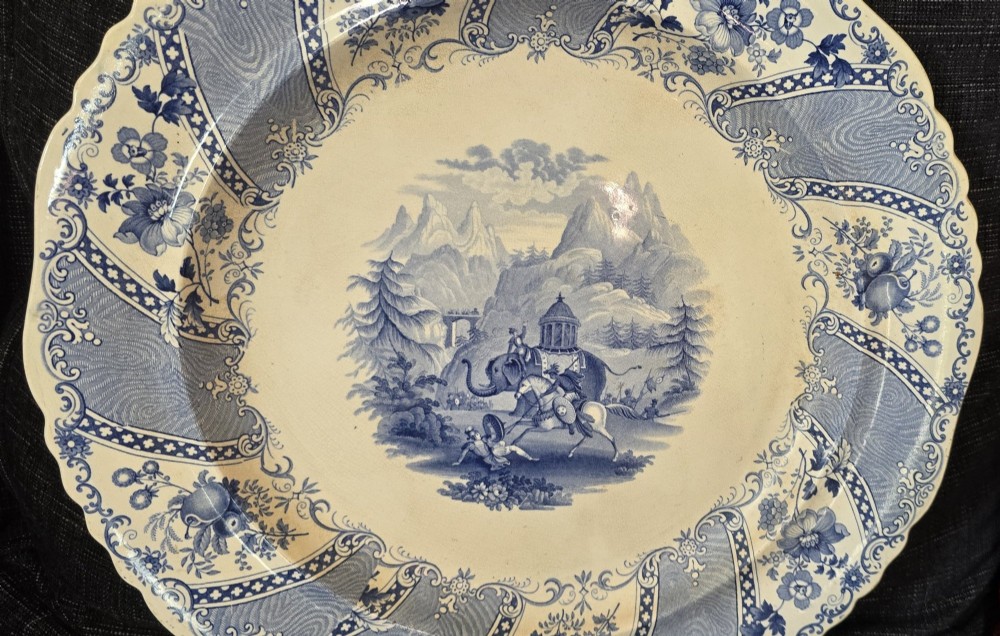 An antique circular earthenware plate featuring a blue transfer-printed central scene of Hannibal with an elephant and figures in a mountainous landscape, surrounded by an ornate scalloped floral border. View II
