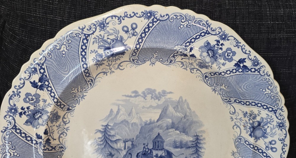 An antique circular earthenware plate featuring a blue transfer-printed central scene of Hannibal with an elephant and figures in a mountainous landscape, surrounded by an ornate scalloped floral border. View IV