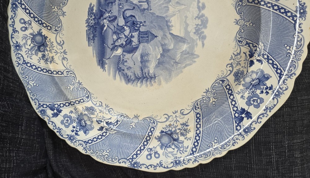 An antique circular earthenware plate featuring a blue transfer-printed central scene of Hannibal with an elephant and figures in a mountainous landscape, surrounded by an ornate scalloped floral border. View VI