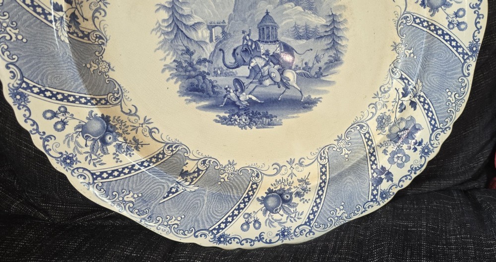 An antique circular earthenware plate featuring a blue transfer-printed central scene of Hannibal with an elephant and figures in a mountainous landscape, surrounded by an ornate scalloped floral border. View VII