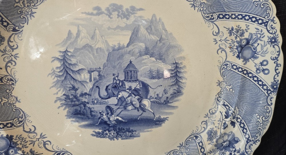 An antique circular earthenware plate featuring a blue transfer-printed central scene of Hannibal with an elephant and figures in a mountainous landscape, surrounded by an ornate scalloped floral border. View VIII