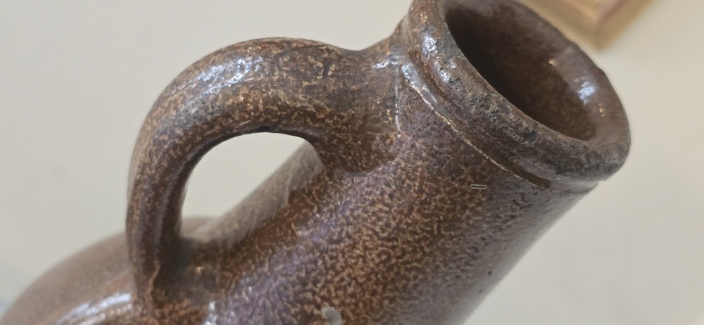 A 17th-century dark brown salt-glazed stoneware bottle featuring a bulbous body, narrow neck, turned base, and a single loop handle. View V
