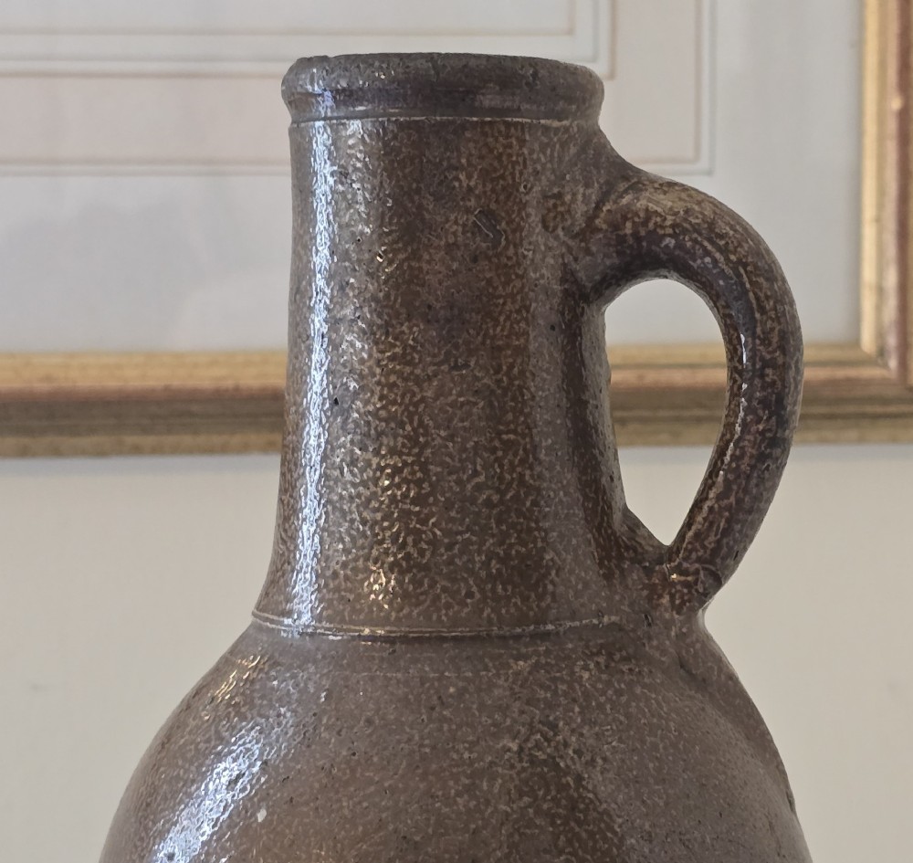 A 17th-century dark brown salt-glazed stoneware bottle featuring a bulbous body, narrow neck, turned base, and a single loop handle. View VI
