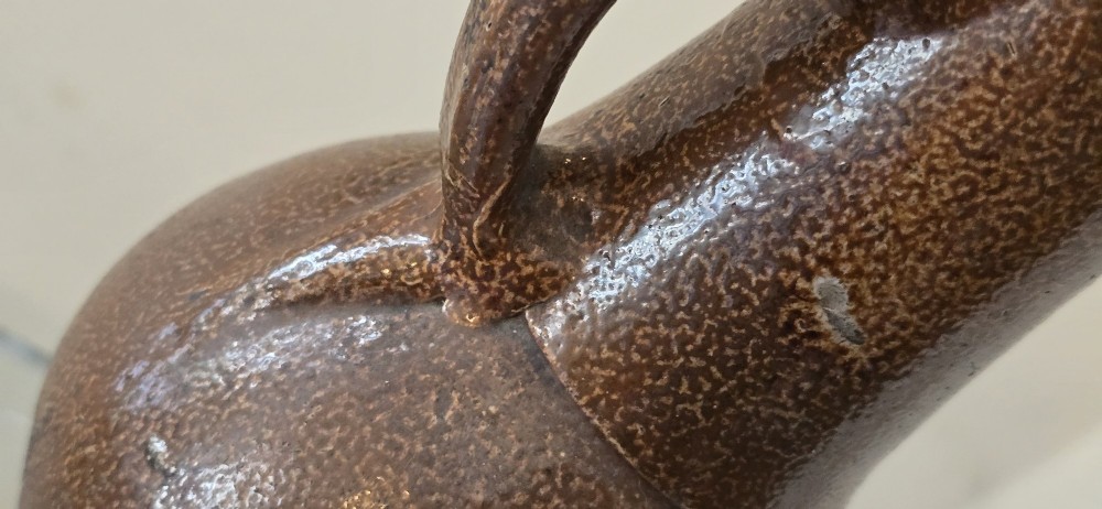 A 17th-century dark brown salt-glazed stoneware bottle featuring a bulbous body, narrow neck, turned base, and a single loop handle. View IX