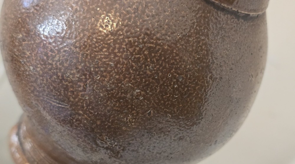 A 17th-century dark brown salt-glazed stoneware bottle featuring a bulbous body, narrow neck, turned base, and a single loop handle. View X