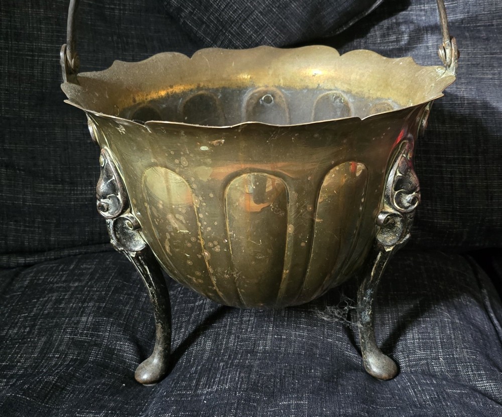 An Arts and Crafts brass coal scuttle featuring a scalloped rim, vertical body fluting, three dark cast legs, and an overhead coiled handle. View II