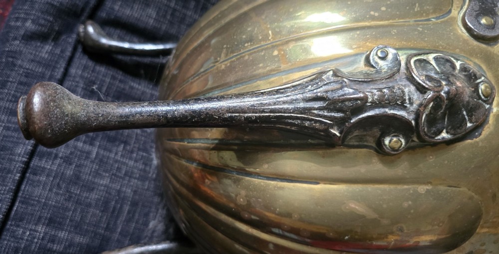 An Arts and Crafts brass coal scuttle featuring a scalloped rim, vertical body fluting, three dark cast legs, and an overhead coiled handle. View VI