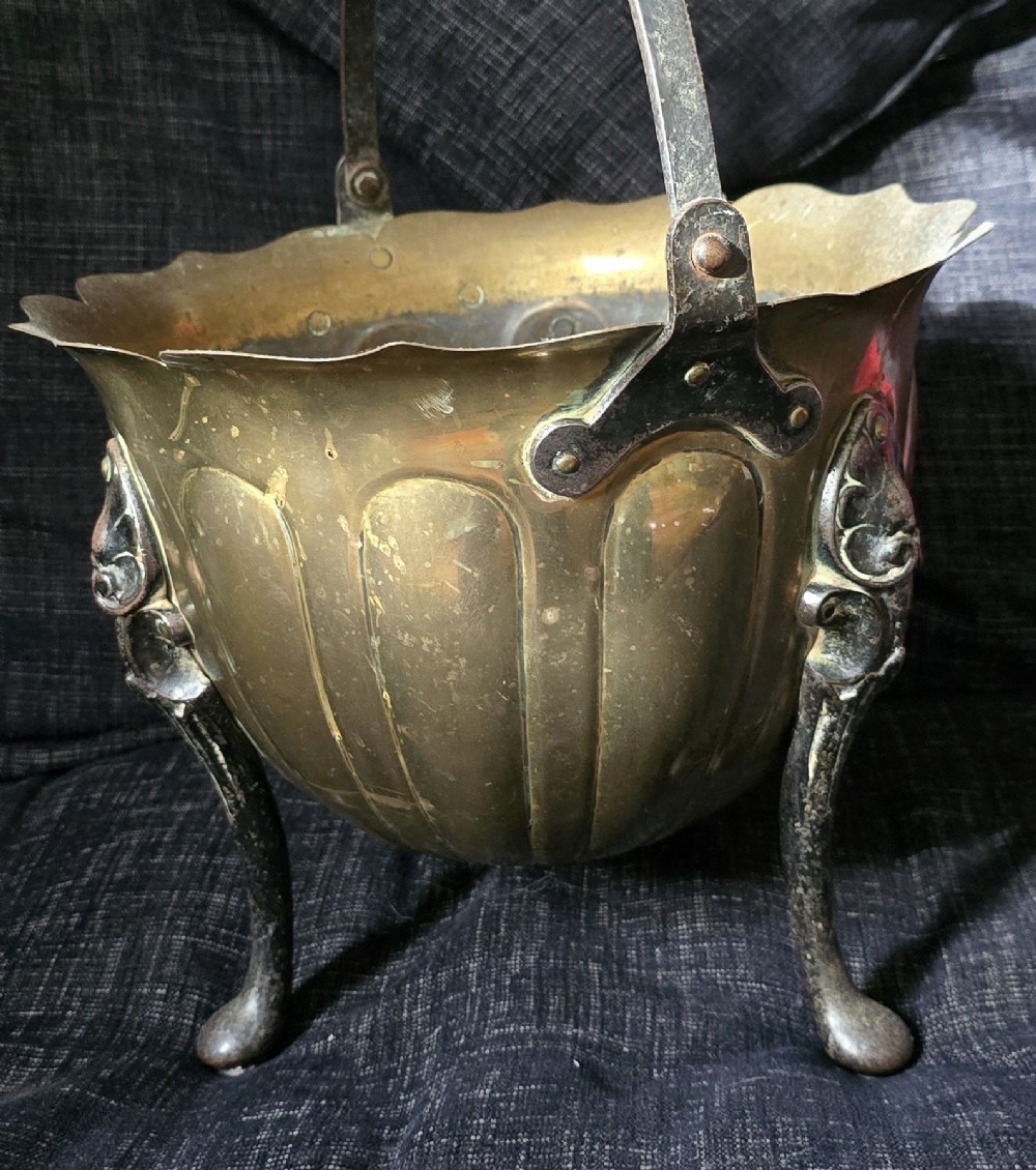 An Arts and Crafts brass coal scuttle featuring a scalloped rim, vertical body fluting, three dark cast legs, and an overhead coiled handle. View X