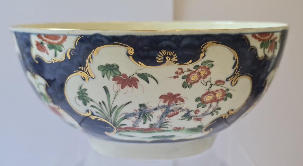 An eighteenth-century circular porcelain bowl featuring a blue scale ground, gilded rococo cartouches, and hand-painted Kakiemon floral panels.