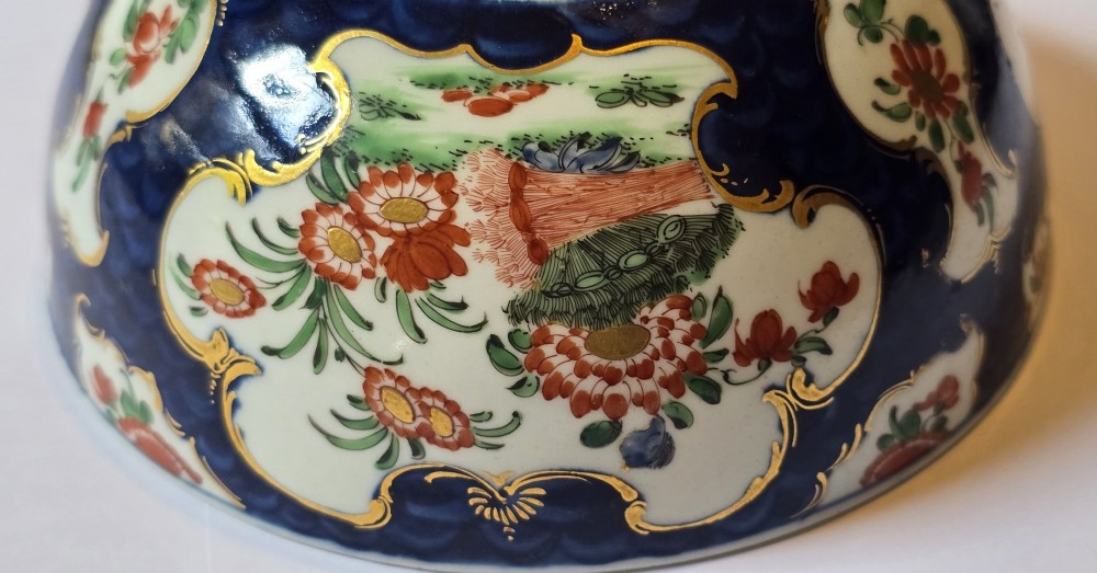 An eighteenth-century circular porcelain bowl featuring a blue scale ground, gilded rococo cartouches, and hand-painted Kakiemon floral panels. View X