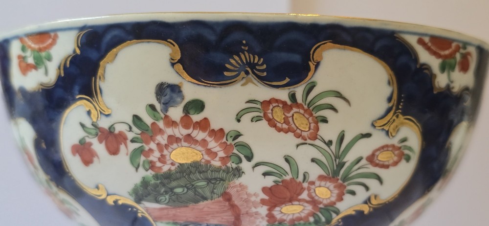 An eighteenth-century circular porcelain bowl featuring a blue scale ground, gilded rococo cartouches, and hand-painted Kakiemon floral panels. View XII
