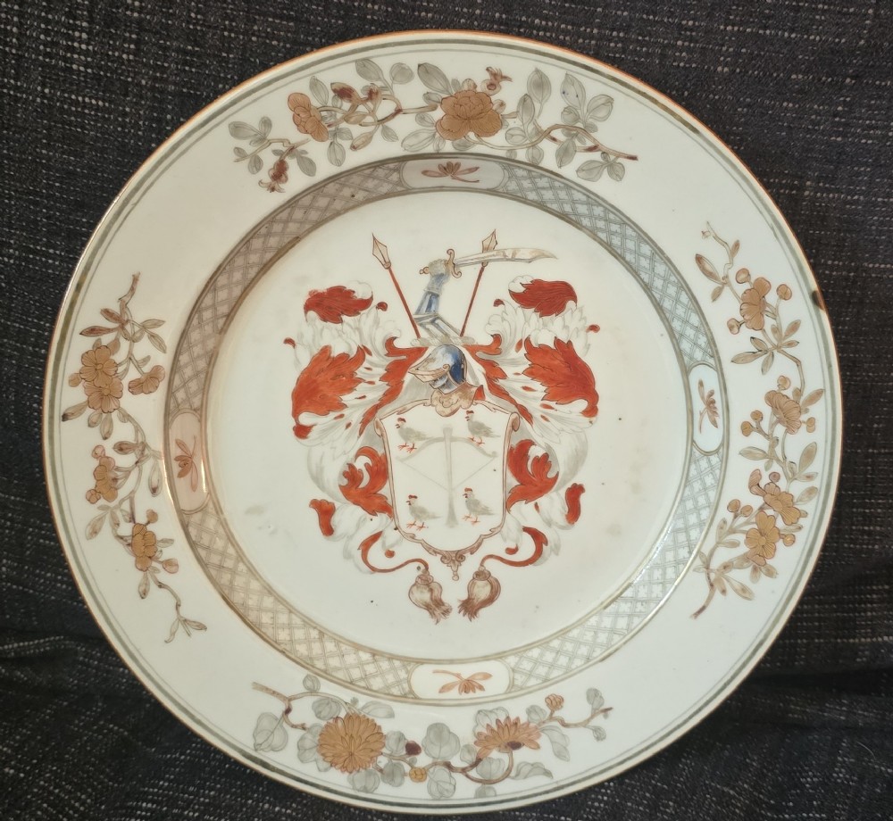 A circular Chinese export porcelain plate featuring a central heraldic crest with red mantling, encircled by a geometric diaper border and floral sprigs.
