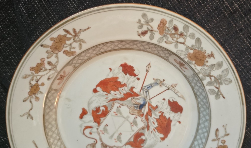 A circular Chinese export porcelain plate featuring a central heraldic crest with red mantling, encircled by a geometric diaper border and floral sprigs. View V