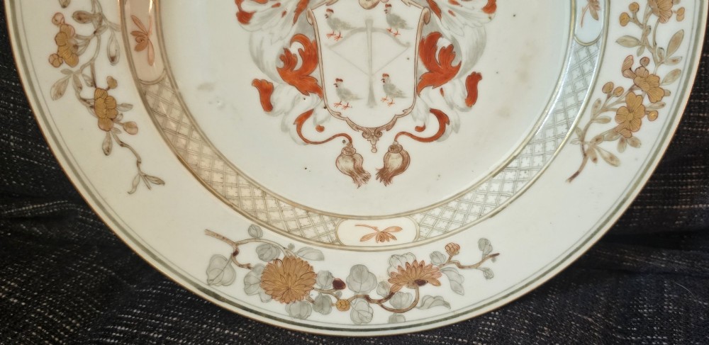 A circular Chinese export porcelain plate featuring a central heraldic crest with red mantling, encircled by a geometric diaper border and floral sprigs. View VI