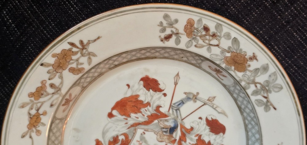 A circular Chinese export porcelain plate featuring a central heraldic crest with red mantling, encircled by a geometric diaper border and floral sprigs. View VII