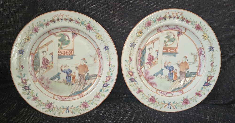 A pair of circular white porcelain plates hand-painted with famille rose enamels depicting figures in an interior and garden setting with a floral rim border.