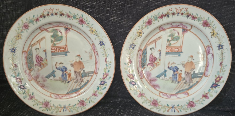 A pair of circular white porcelain plates hand-painted with famille rose enamels depicting figures in an interior and garden setting with a floral rim border. View II