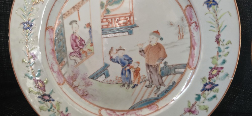 A pair of circular white porcelain plates hand-painted with famille rose enamels depicting figures in an interior and garden setting with a floral rim border. View VI