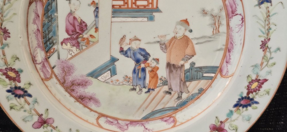 A pair of circular white porcelain plates hand-painted with famille rose enamels depicting figures in an interior and garden setting with a floral rim border. View VII