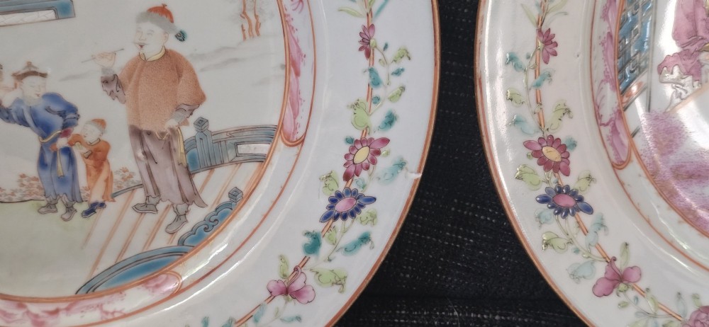 A pair of circular white porcelain plates hand-painted with famille rose enamels depicting figures in an interior and garden setting with a floral rim border. View XIV