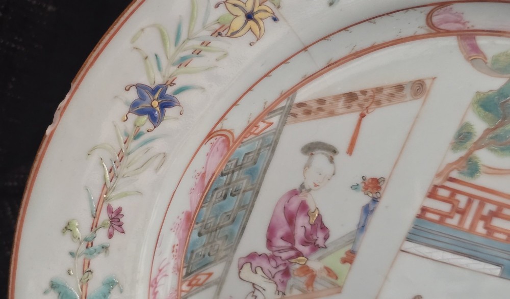 A pair of circular white porcelain plates hand-painted with famille rose enamels depicting figures in an interior and garden setting with a floral rim border. View XV