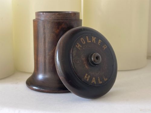 fig 1. A small turned dark wooden go-to-bed match holder featuring a cylindrical body, a moulded cap, and a match inserted in the centre top.
