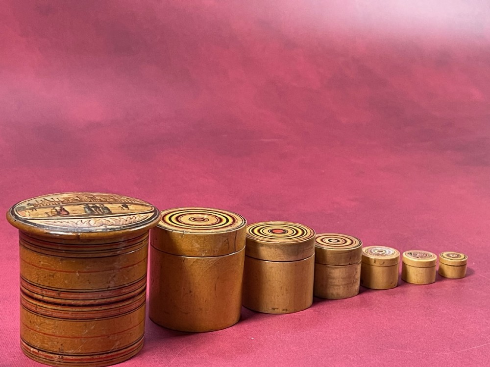 A graduated set of seven cylindrical turned whitewood stacking pots featuring a printed landscape view on the largest lid and concentric coloured circles on the smaller lids. View II