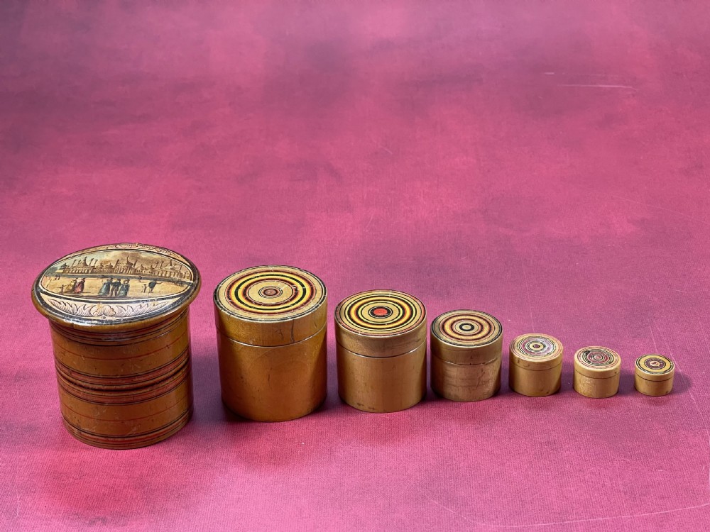 A graduated set of seven cylindrical turned whitewood stacking pots featuring a printed landscape view on the largest lid and concentric coloured circles on the smaller lids. View V