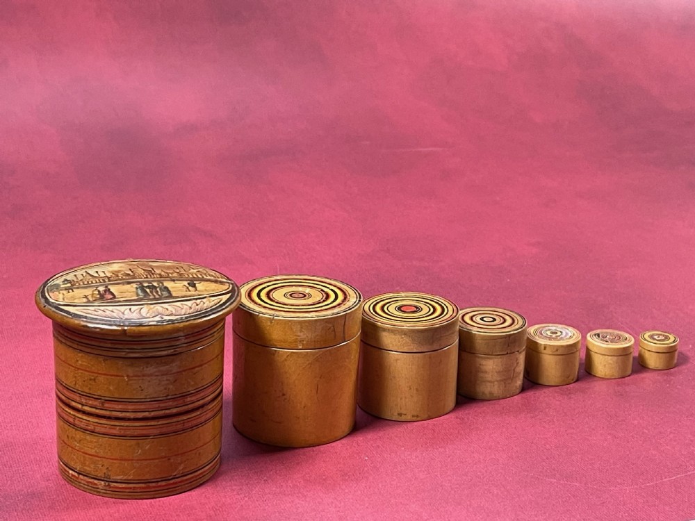 A graduated set of seven cylindrical turned whitewood stacking pots featuring a printed landscape view on the largest lid and concentric coloured circles on the smaller lids. View VI