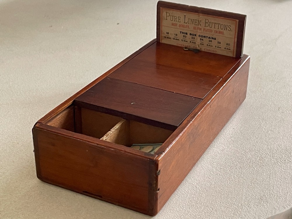 A rectangular wooden haberdashery display box featuring sliding lid panels and an upright backboard holding a printed pure linen buttons label.