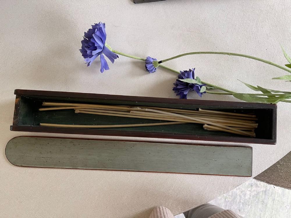 An open antique wooden taper box displaying several thin rushlight tapers inside alongside its separate lid.