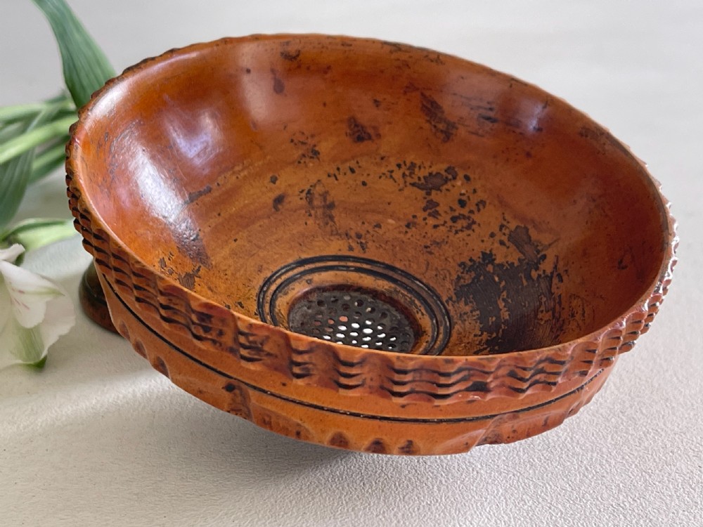 A small turned warm brown wooden Georgian searce with a carved wavy rim, pedestal base, and perforated strainer bottom. View V