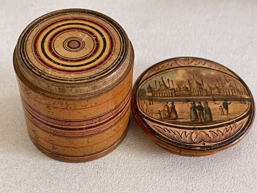 A set of seven turned natural wood nesting pots featuring colourful concentric painted rings on the lids and a printed landscape on the largest cover. View II