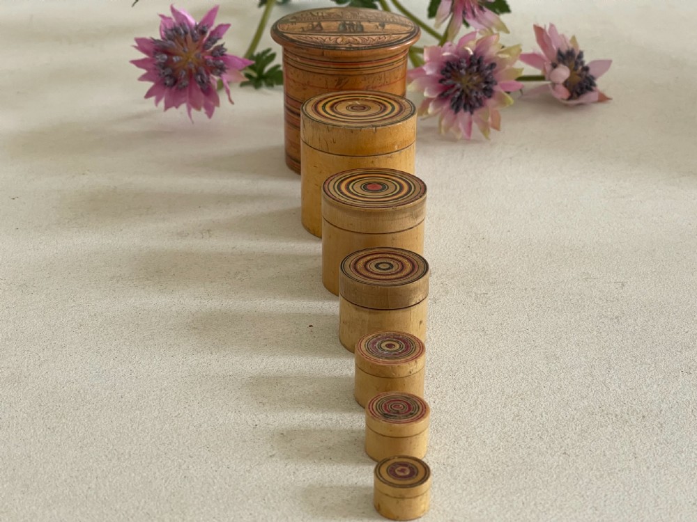 A set of seven turned natural wood nesting pots featuring colourful concentric painted rings on the lids and a printed landscape on the largest cover. View IV